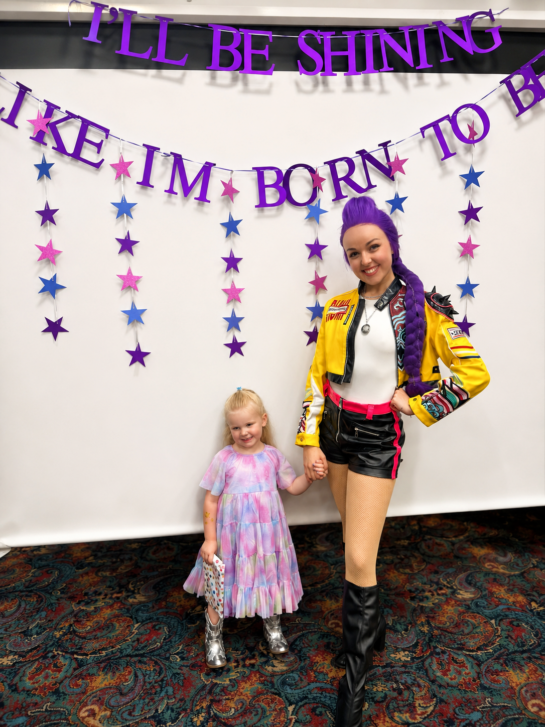 A woman with purple hair and a young girl holding hands, standing in front of a backdrop with purple and pink star decorations and a sign that reads 'I'll be shining like I'm born to be'.