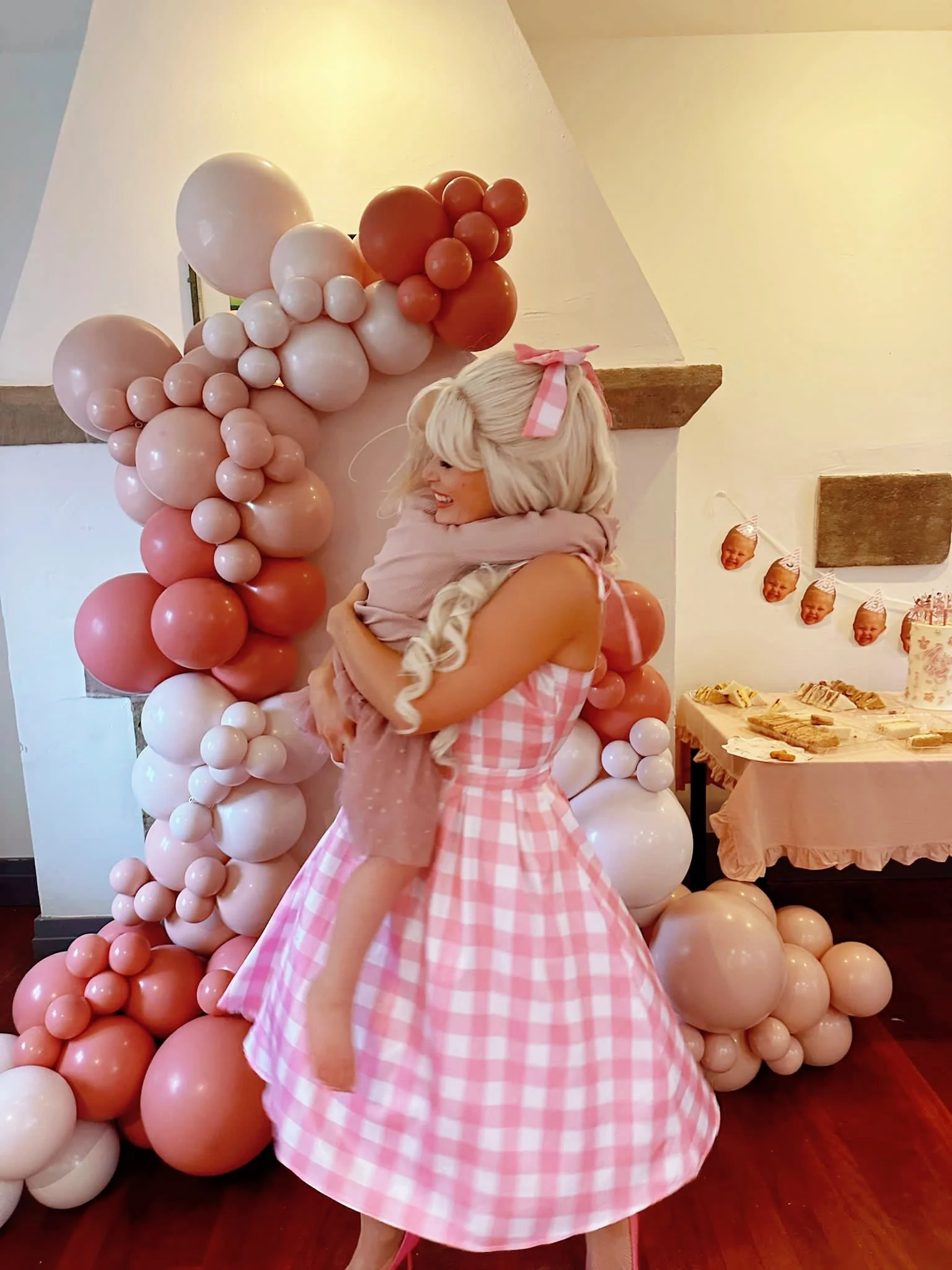 A woman dressed in a pink and white checkered party dress hugging a child at a celebration with pink, white, and peach balloons and party decorations.