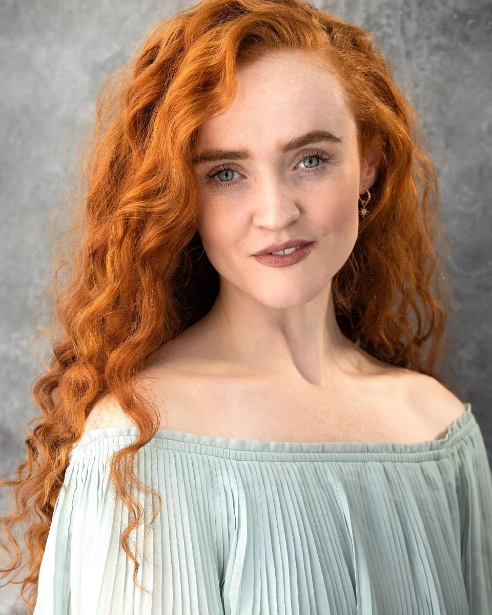 A young woman with long, curly red hair, fair skin, and blue eyes, wearing a light green off-the-shoulder top, earrings, and natural makeup, standing against a gray background.