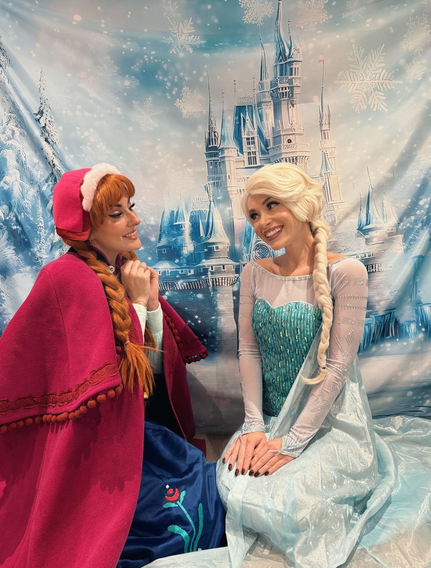 Two women dressed as Disney characters Elsa and Anna from Frozen, sitting and smiling at each other in front of a castle backdrop with snowflakes.