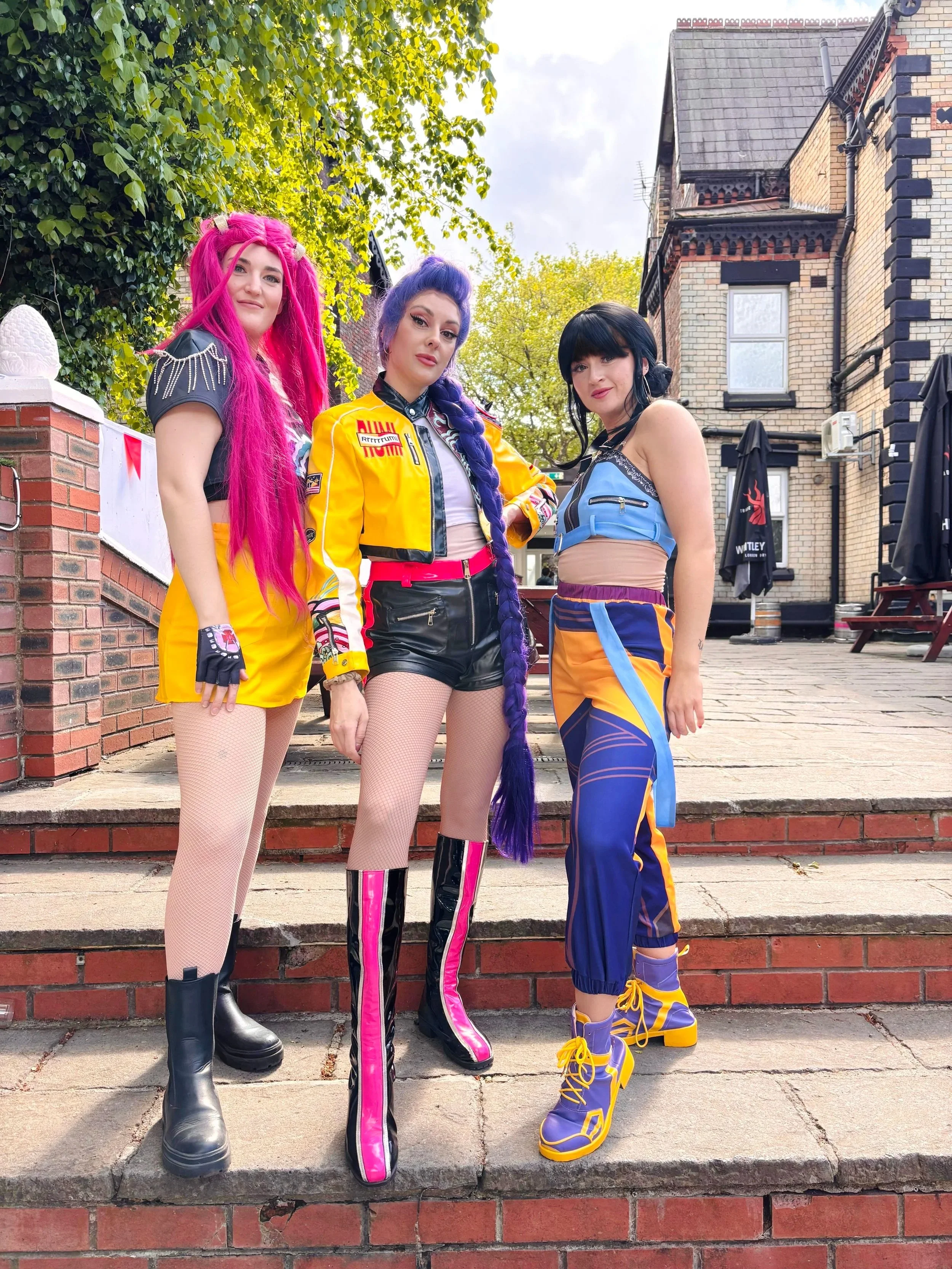 Three women with colorful hair and outfits standing on a brick sidewalk in front of a brick building with umbrellas and trees in the background.