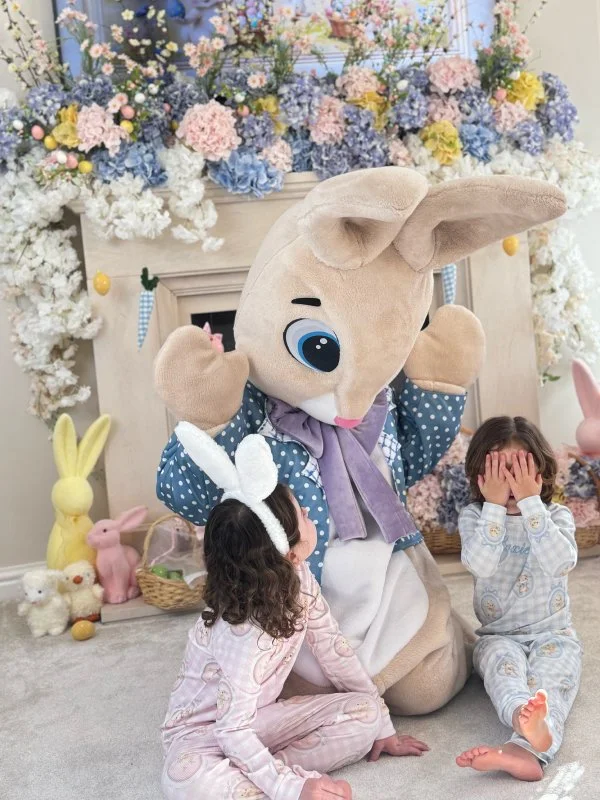 Costumed character of a large beige bunny with a gray scarf, surrounded by two children in pajamas and Easter decorations including pastel-colored flowers, bunny figurines, and plush toys.