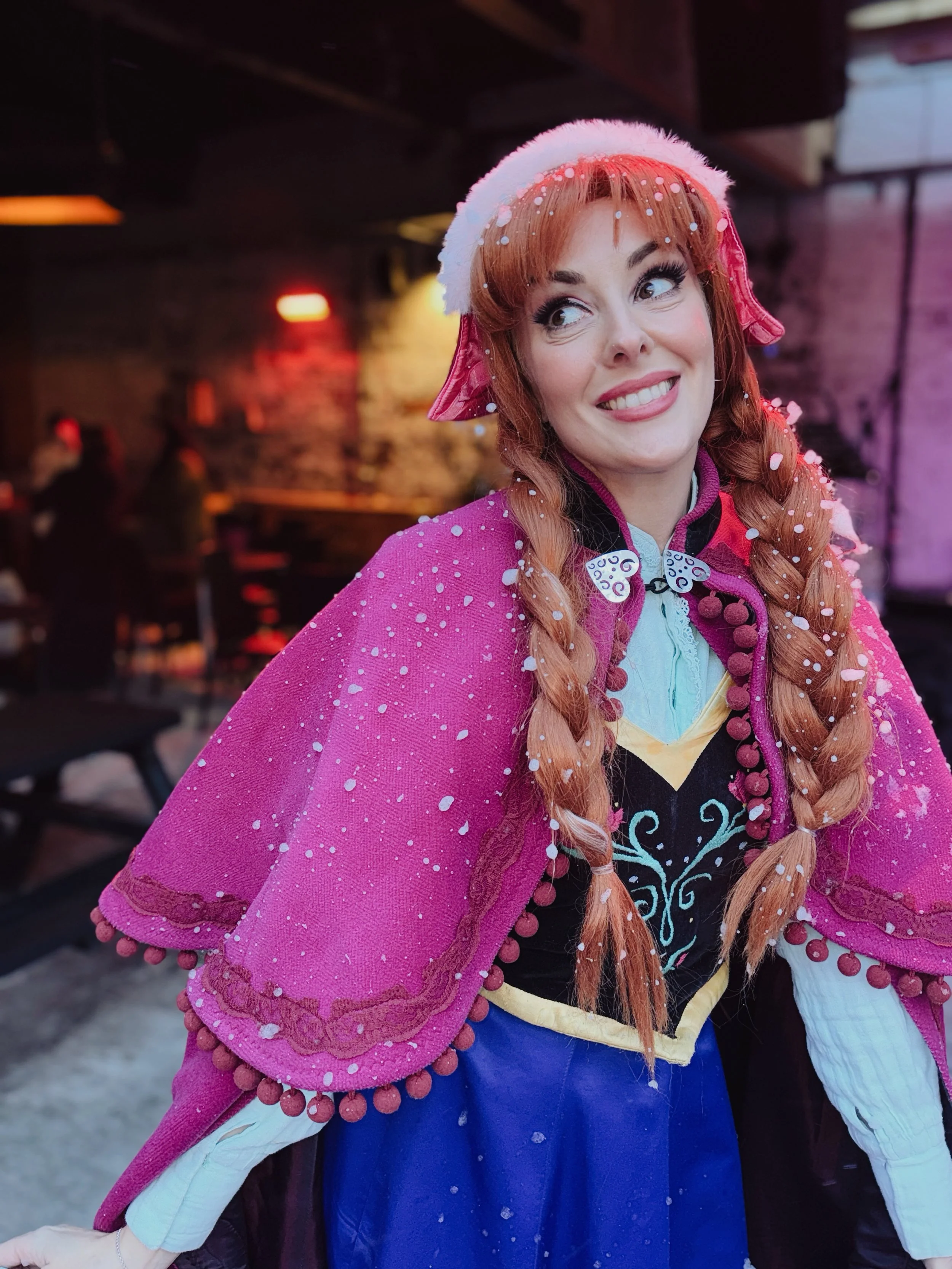 A woman dressed as Anna from Frozen, wearing a pink cape with pom-pom trim, a pink beret, and a dress with blue and white details.