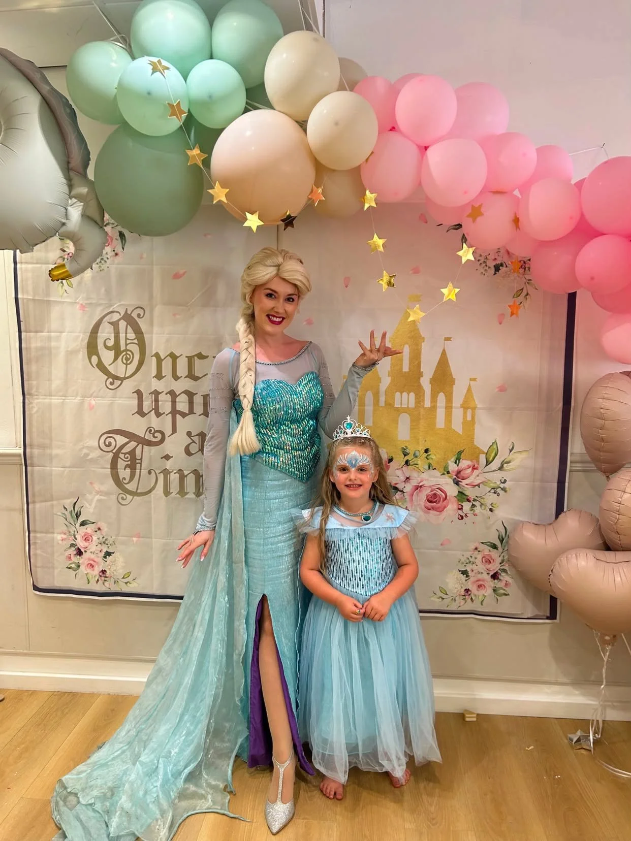 A woman dressed as Elsa from Frozen and a young girl dressed as Elsa standing in front of a castle backdrop with pink and green balloons and star-shaped decorations at a themed birthday party.