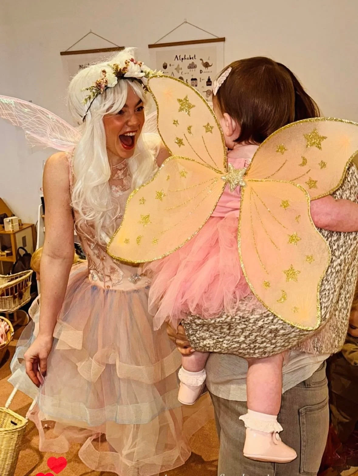 Child in pink fairy costume with large glittery wings being held by an adult, woman dressed as a fairy with white hair, fairy wings, and a floral crown, smiling and interacting in a room decorated with posters and baskets.