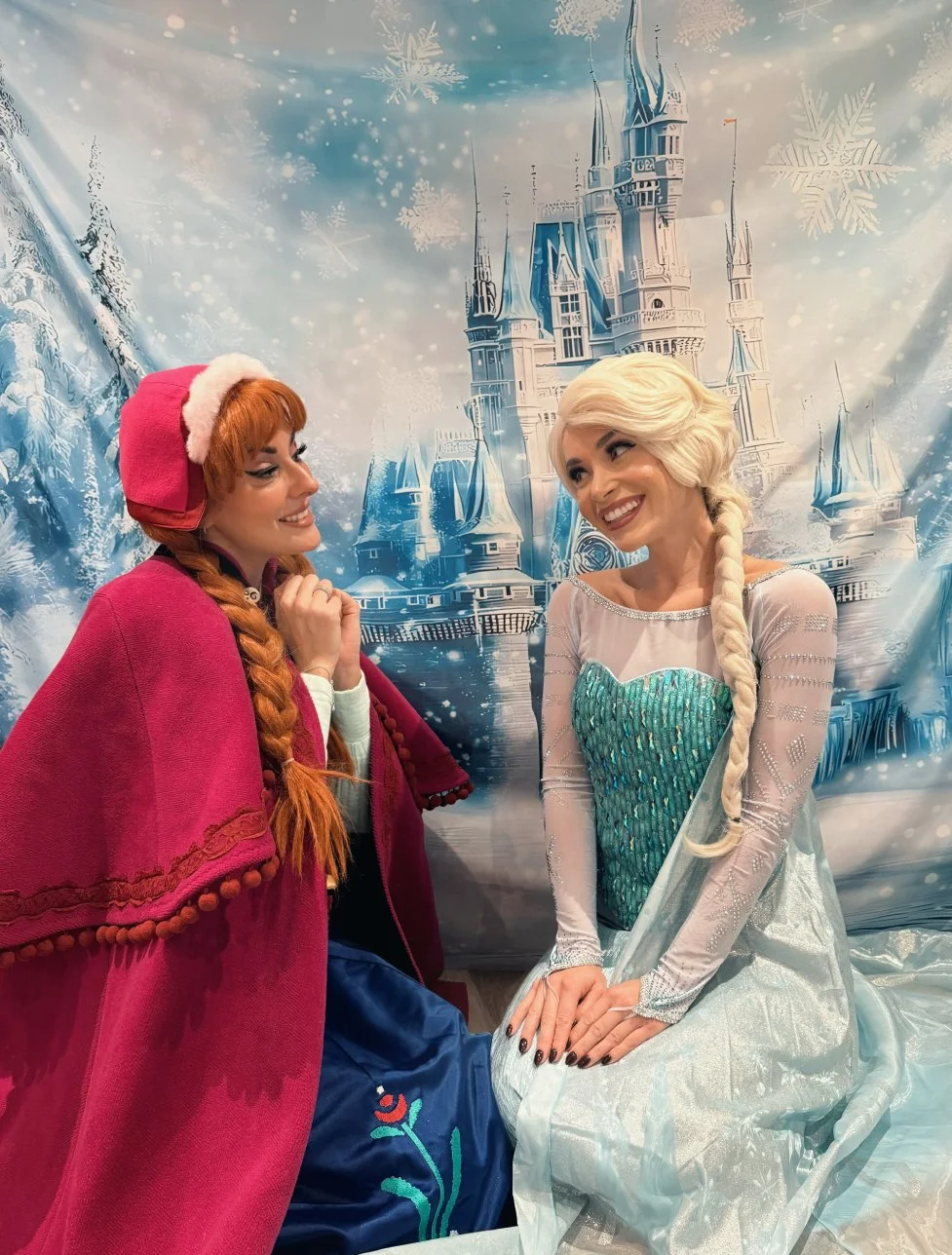 Two women dressed as Disney characters Elsa and Anna from Frozen, smiling at each other, with a castle and wintery scene backdrop.