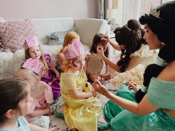 Group of young girls in princess costumes and crowns sitting on a sofa, while a woman dressed as a princess applies makeup to one of the girls.