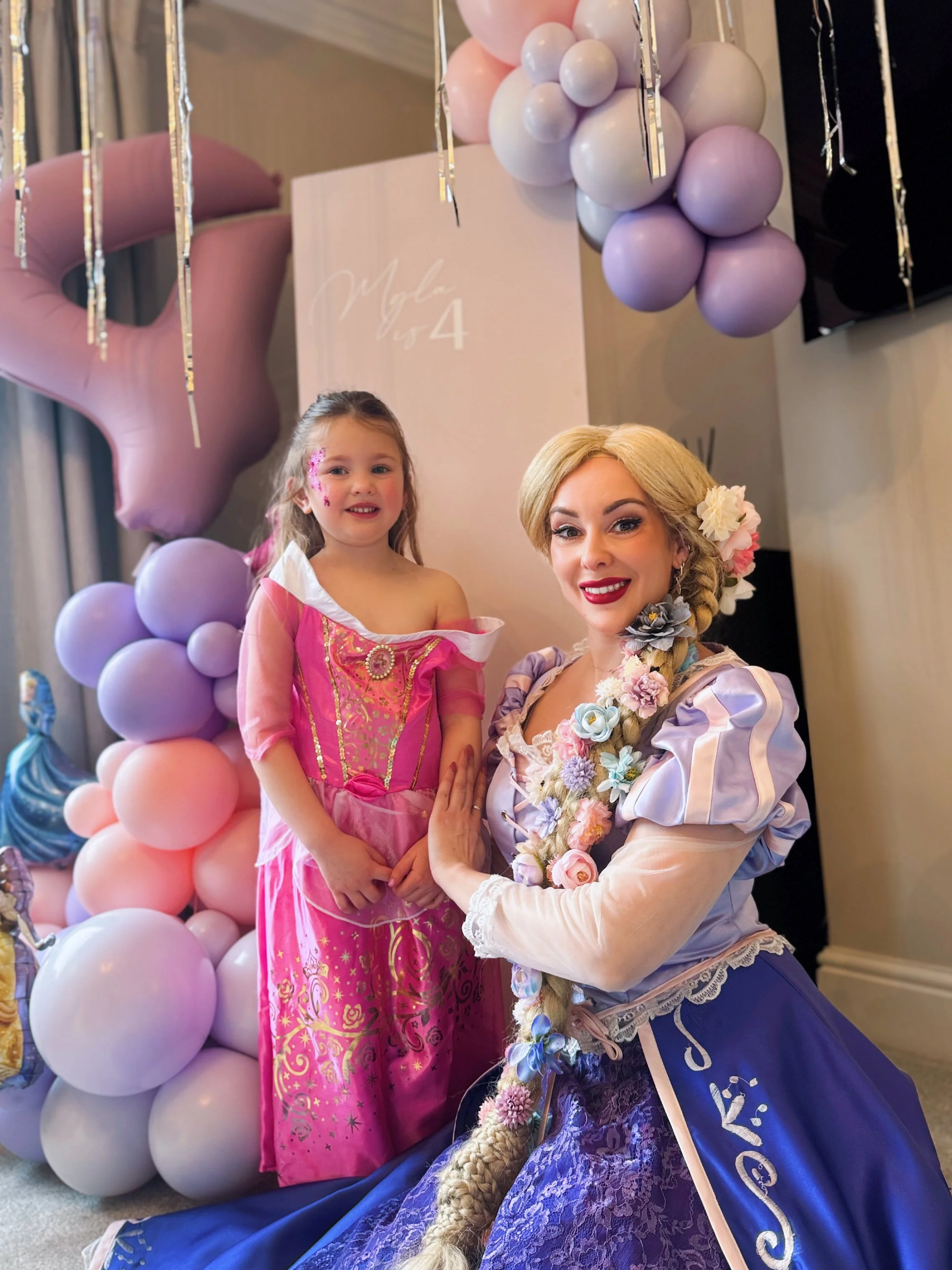 A woman with blonde hair and floral decorations in her hair is dressed as a princess in a blue gown with a pink sash, smiling with a young girl dressed as a princess in a pink costume. They are posing in front of a balloon arrangement with pink, purp