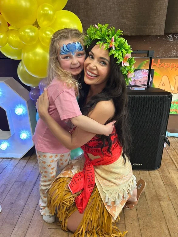 A young girl with face paint hugging a woman dressed in a lei, a grass skirt, and a floral headpiece, at a party with yellow balloons and a festive backdrop.
