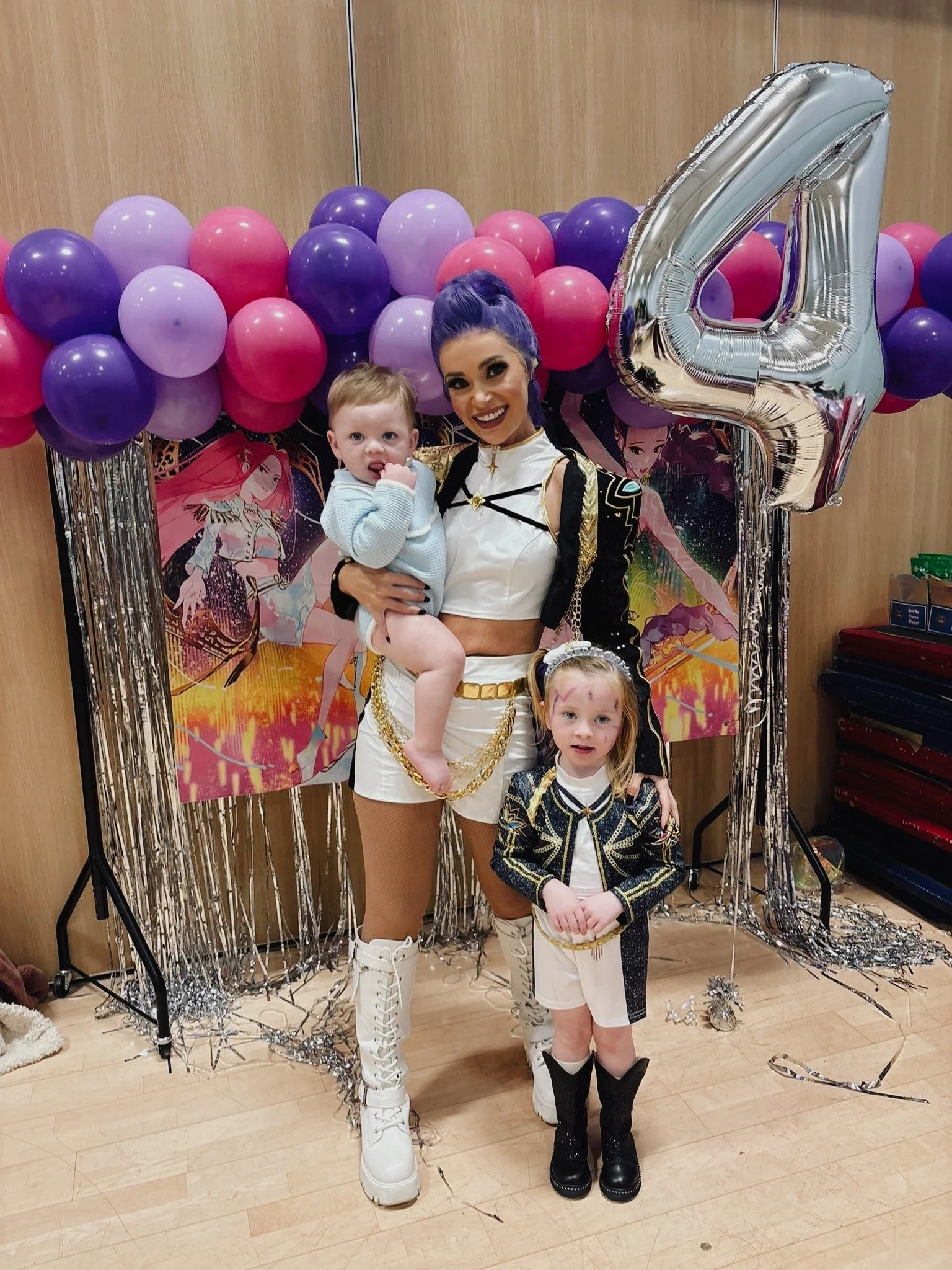 A woman and three children dressed in stylish outfits stand in front of colorful balloons and a poster with animated characters. The woman wears a white crop top, gold chains, and white boots. The children are in black and gold outfits, with the girl