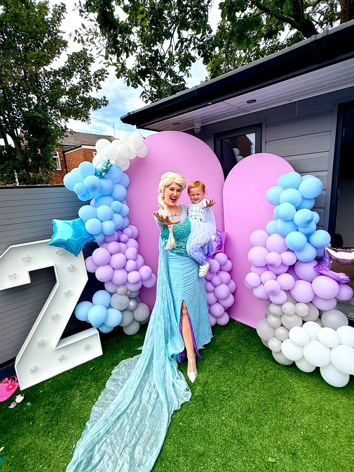 A woman dressed as Elsa from Frozen holding a young girl at a birthday celebration. They are standing in front of a pink backdrop decorated with balloons in shades of purple, blue, and white, with a large white number 2 light-up decoration.
