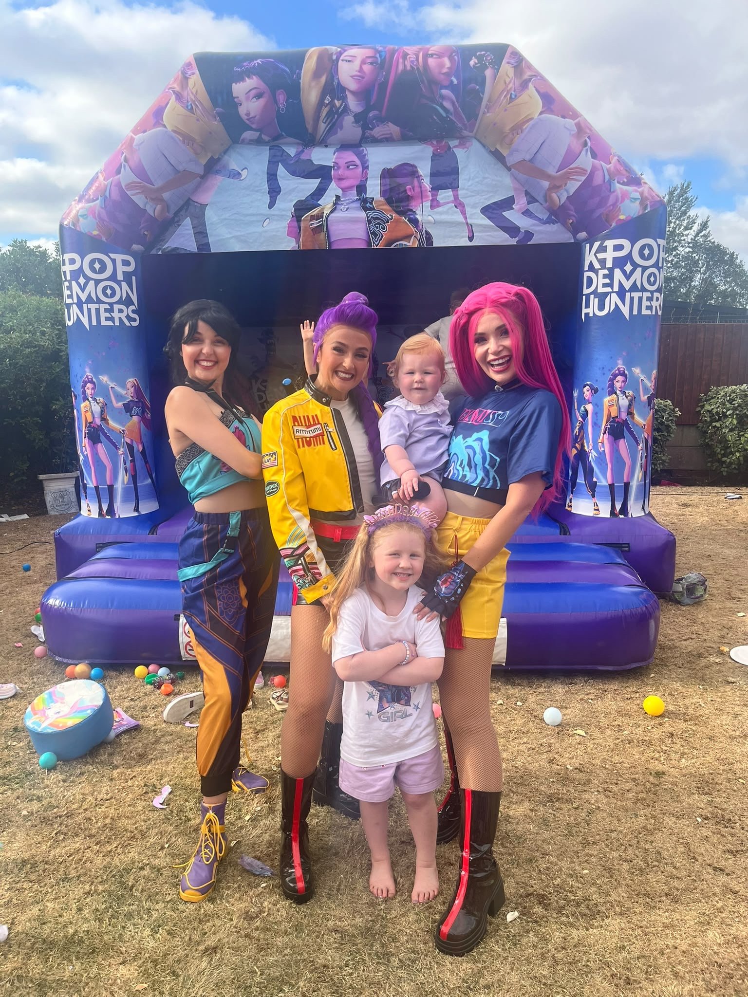 Four women and two children standing in front of a large inflatable bounce house themed with animated characters from the game 'Pop Poker Hunters'. The women are smiling and dressed in colorful casual outfits. One woman has pink hair, another has pur