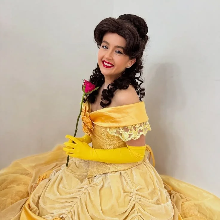 A woman dressed as Belle from Beauty and the Beast in a yellow gown, holding a pink rose, smiling, with curly hair and red lipstick.