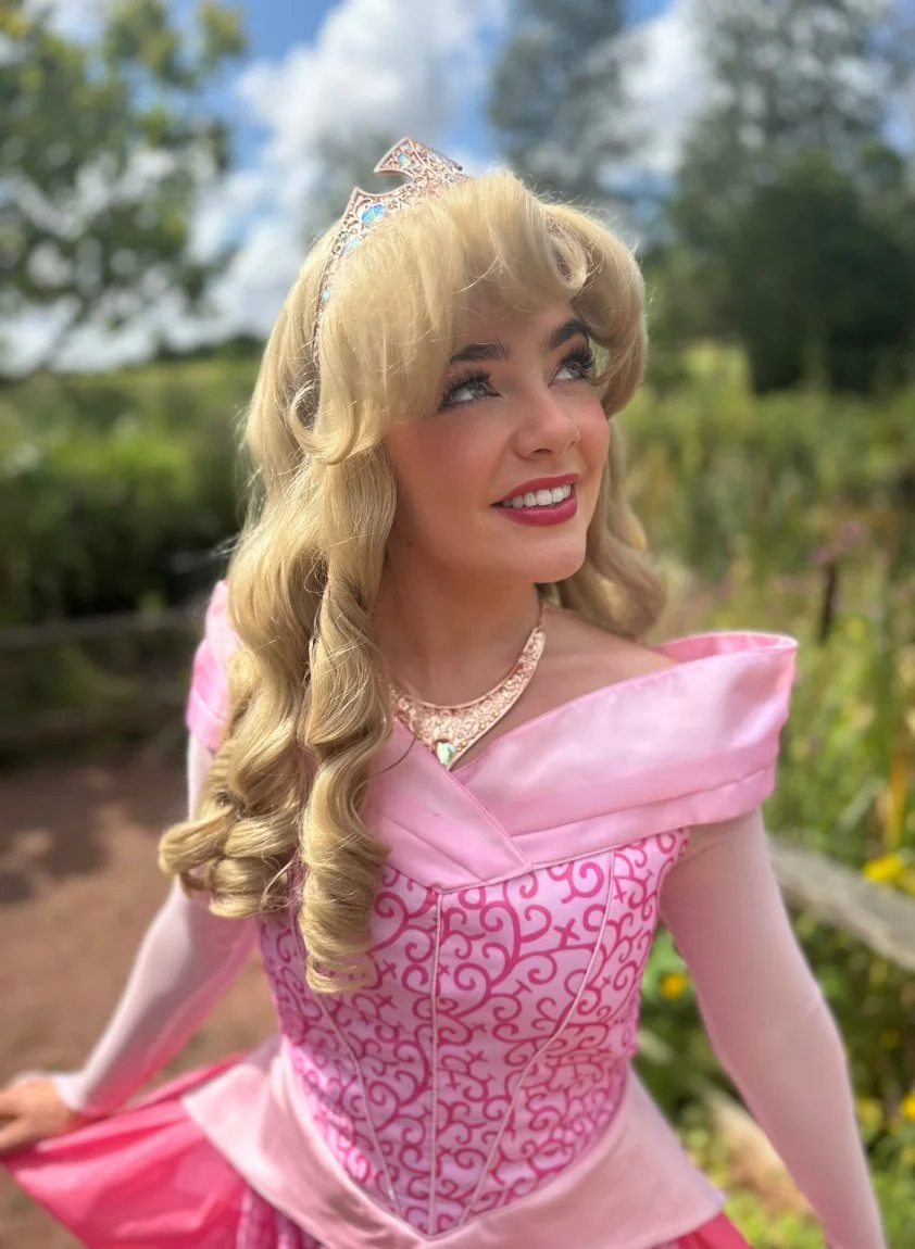 A young woman dressed as a princess, with long curly blonde hair, wearing a pink gown with ornate details, a tiara, and a necklace, smiling and looking upward outdoors with trees in the background.