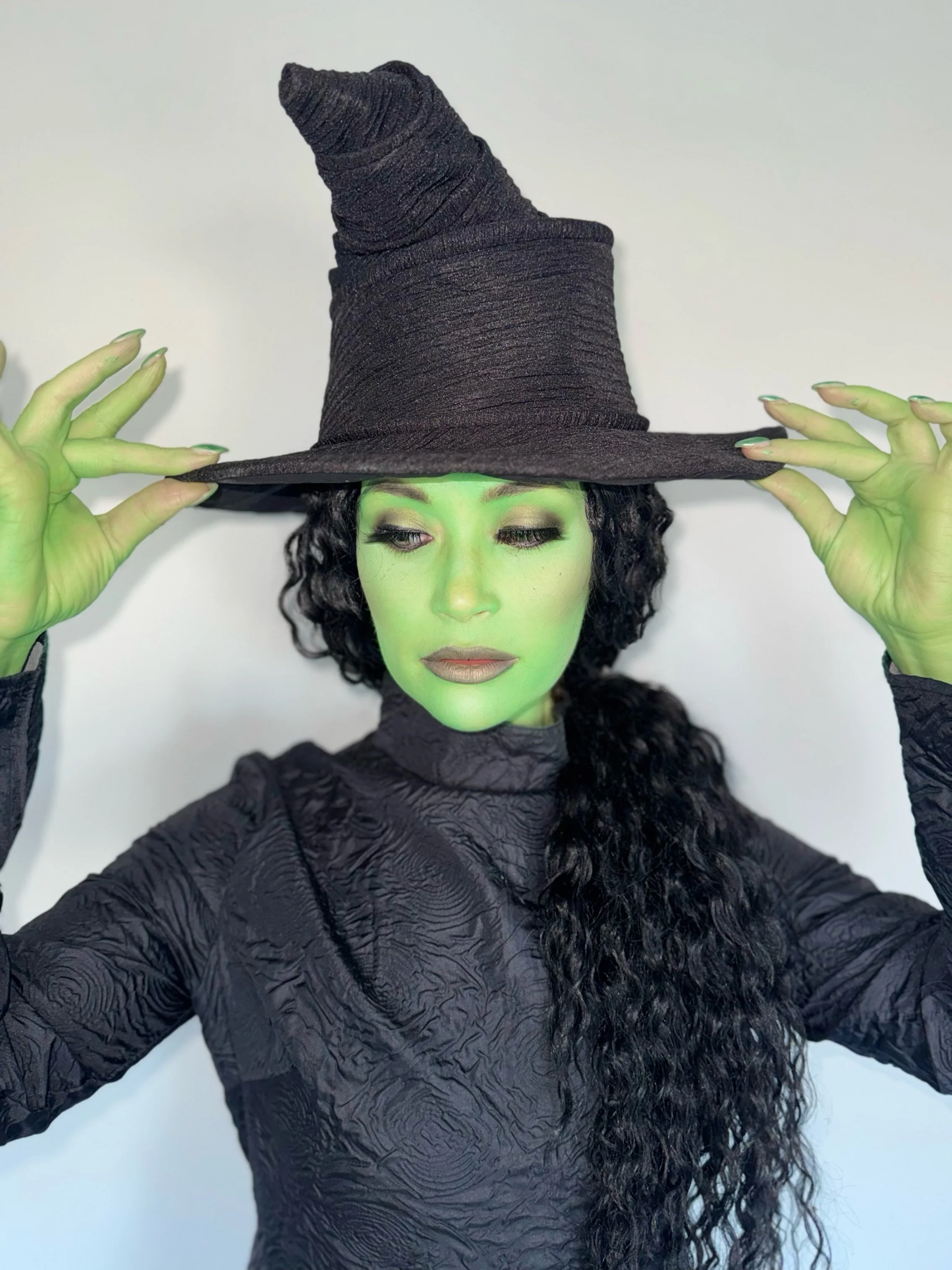 A person dressed as a witch with green face paint, black curly hair, wearing a tall, pointed black witch hat and black textured clothing, holding the brim of the hat with both hands.