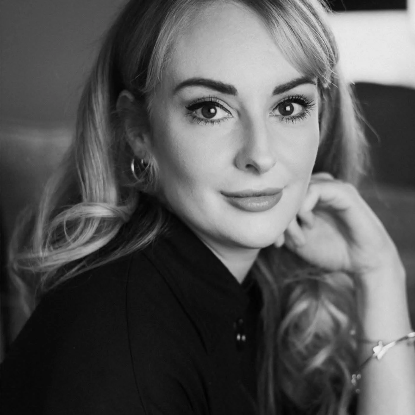 Close-up black and white portrait of a woman with wavy hair, smiling softly, holding her chin with one hand, wearing earrings and a dark top.