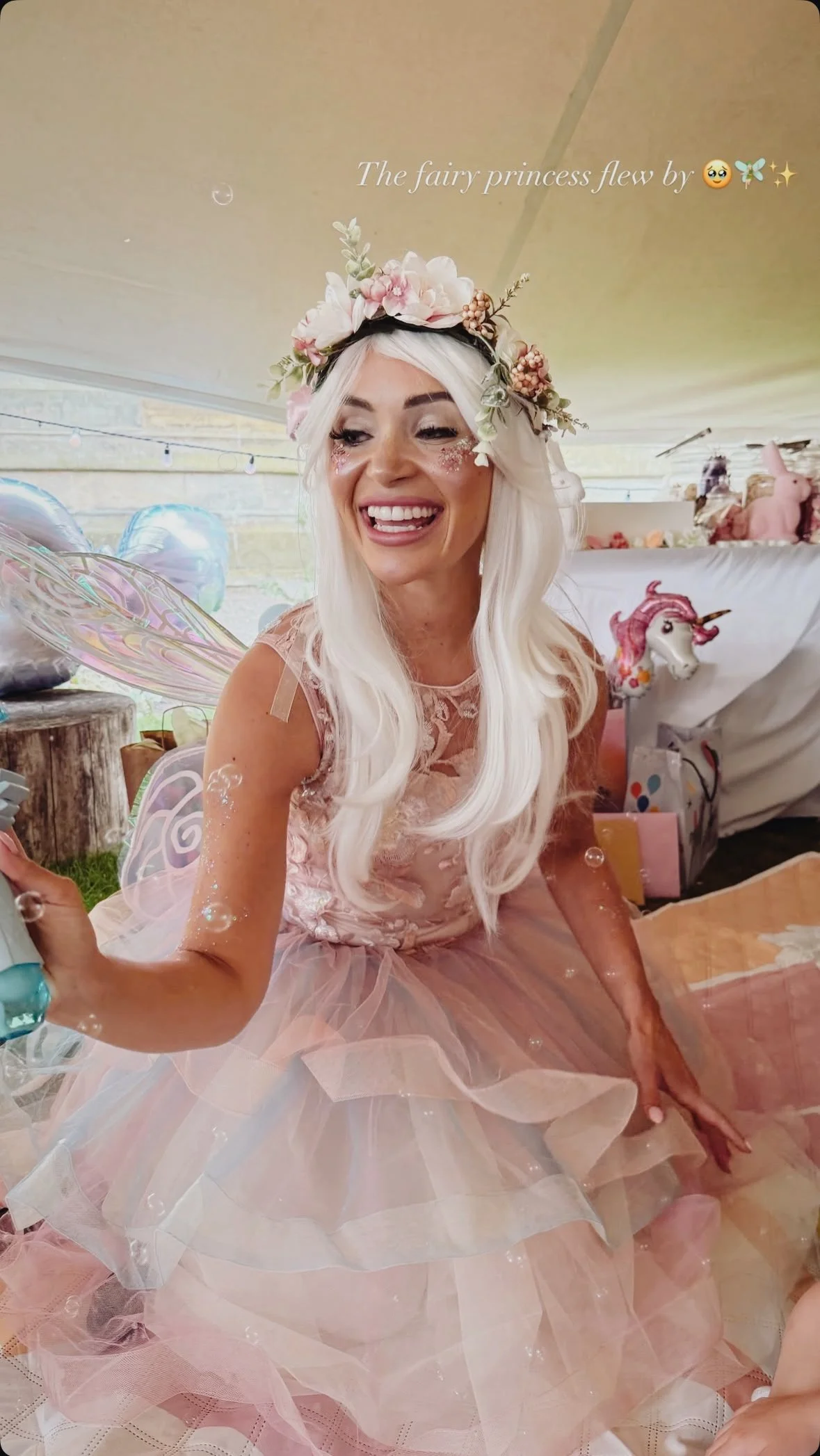 Woman dressed as a fairy princess with long blonde hair, floral crown, pink tulle dress, fairy wings, smiling and surrounded by balloons and party decorations.