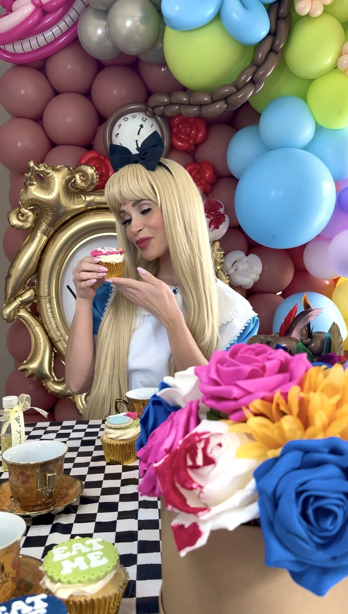 A woman dressed as Alice from Alice in Wonderland, with long blonde hair and a black bow headband, holding a cupcake. She is at a colorful birthday party with balloons, flowers, a checkered tablecloth, and birthday decorations.