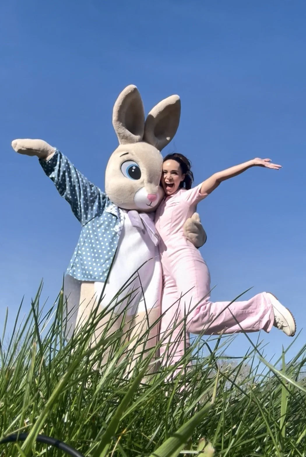 A woman in pink pajamas joyfully jumping in tall grass with a large Easter Bunny mascot dressed in a blue polka dot shirt, against a clear blue sky.