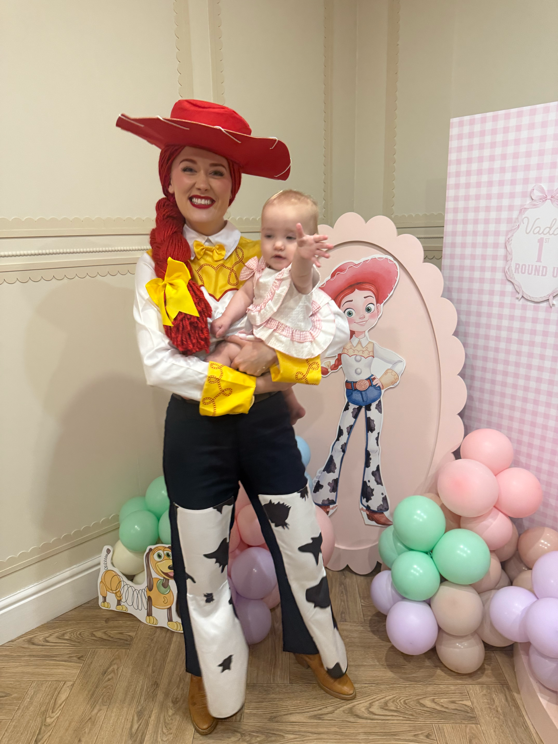 A person dressed as Jessie from Toy Story holding a young child in front of a pink and purple balloon display with a Woody cutout and a sign that says "Vader 1st Round Up" at a birthday party.