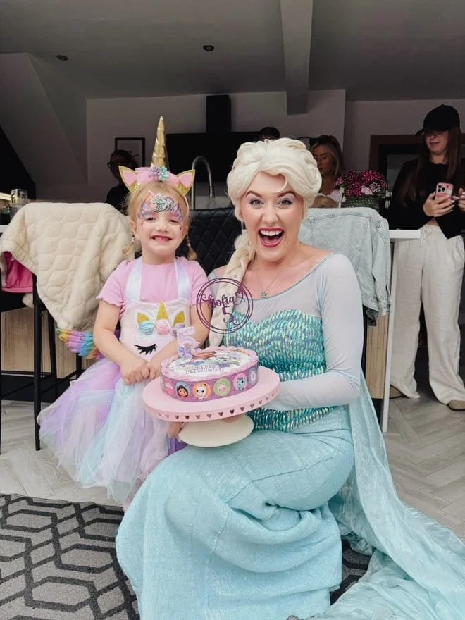 A woman dressed as Elsa from Frozen and a young girl in a unicorn-themed costume celebrating a birthday with a colorful cake on a pink cake stand.