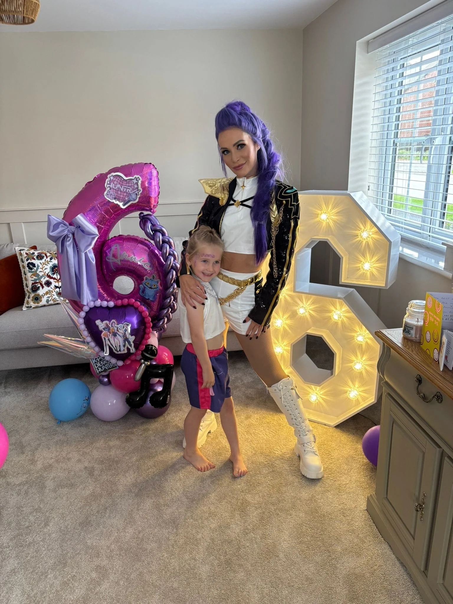 A woman with purple hair in a costume standing next to a young girl with face paint, surrounded by balloons and decorations in a living room.