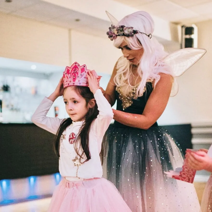 A woman dressed as a fairy with pink hair, fairy wings, and a flower crown helping a young girl put on a pink princess crown at a party.