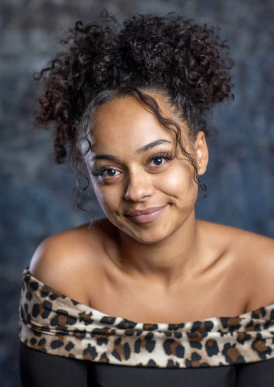 A woman with curly hair and a off-shoulder leopard print top smiling at the camera.