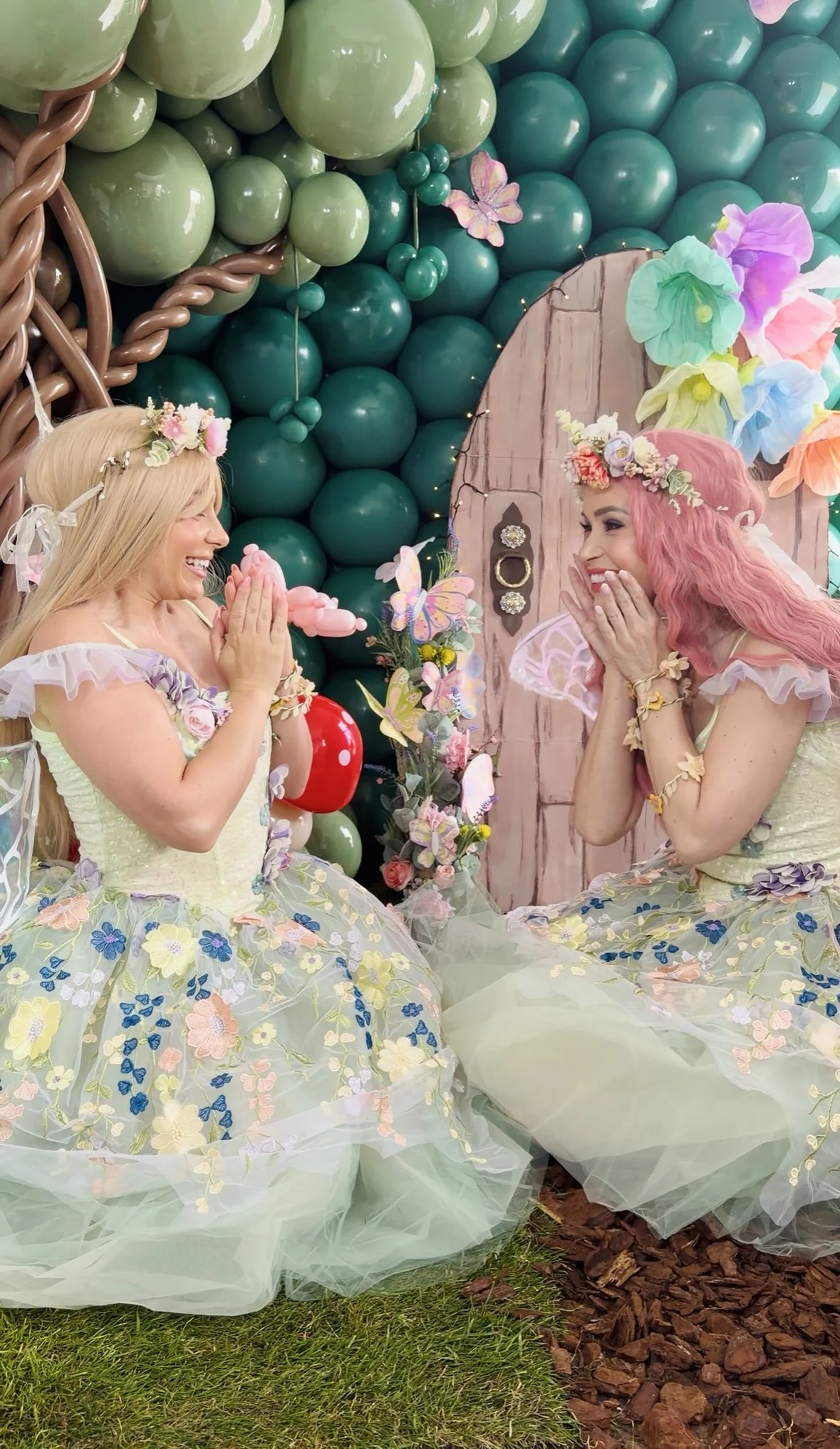 Two women dressed in pastel-colored floral dresses and floral crowns sit facing each other, smiling with their hands near their faces in front of a decorative background of green balloons, colorful paper flowers, butterflies, and fairy lights.