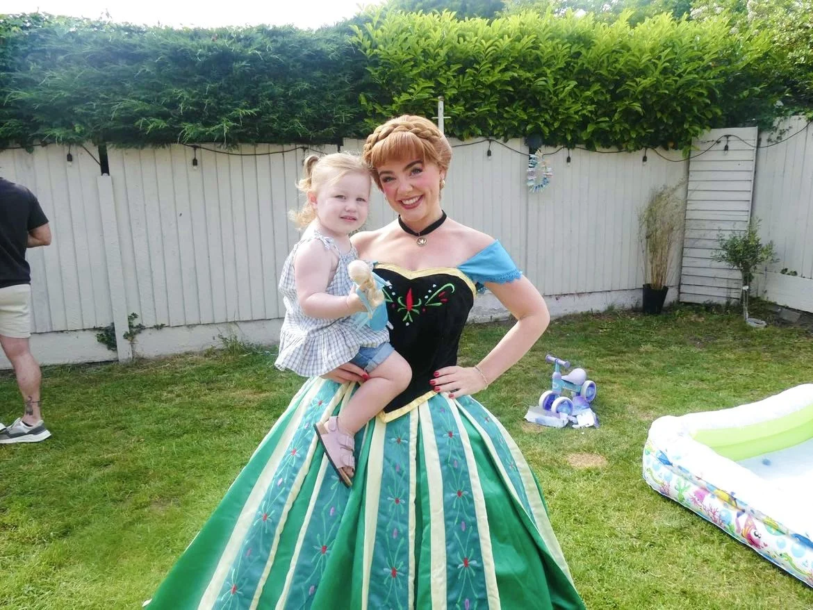 A woman dressed as a princess holding a young girl in a backyard garden with a white fence, greenery, a mini pool, and toys in the background.