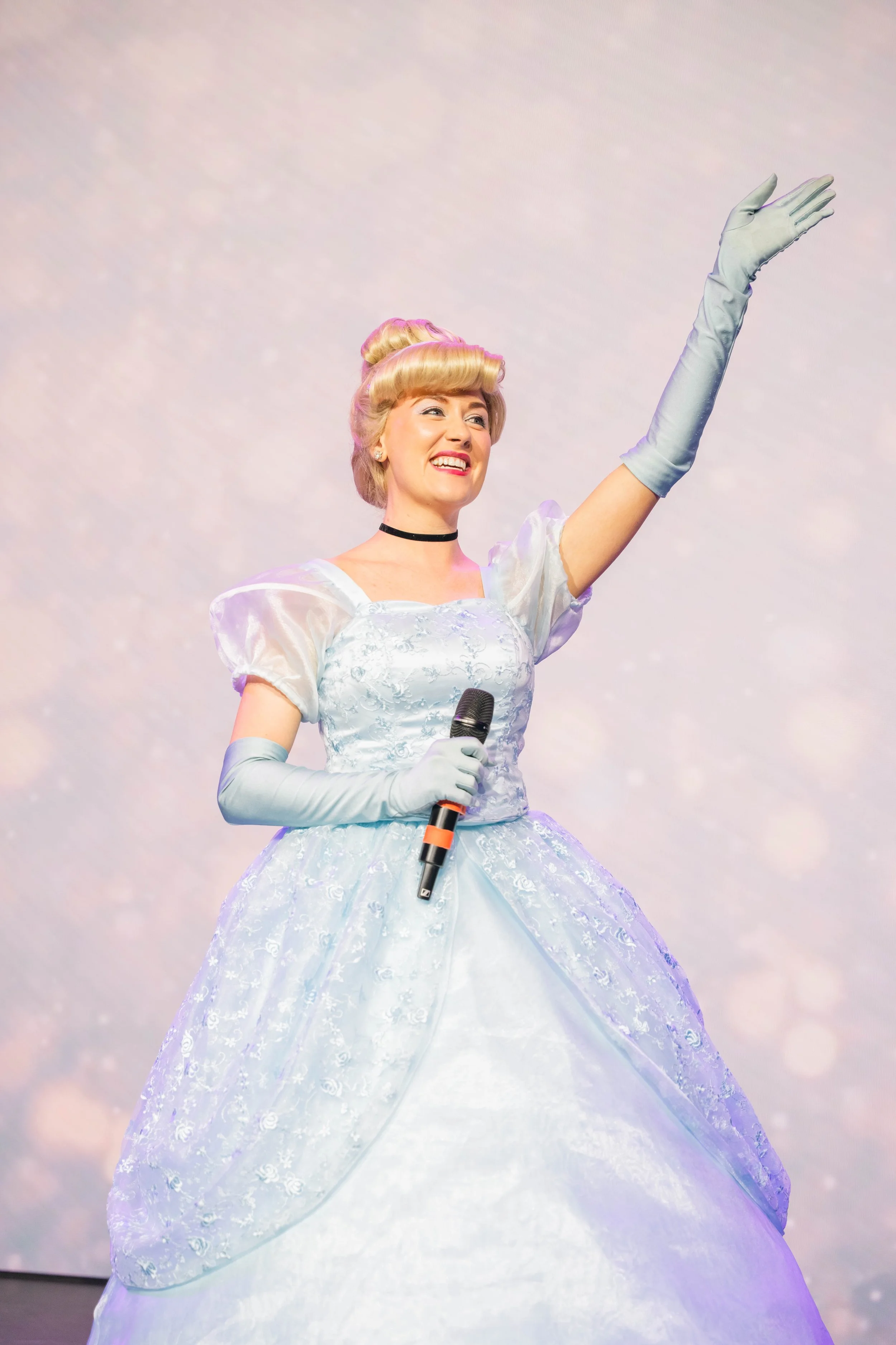 A woman dressed as Cinderella, wearing a light blue ball gown with lace details, long gloves, and a choker, smiling and waving with her right hand while holding a microphone in her left hand, standing against a sparkly, pastel-colored background.