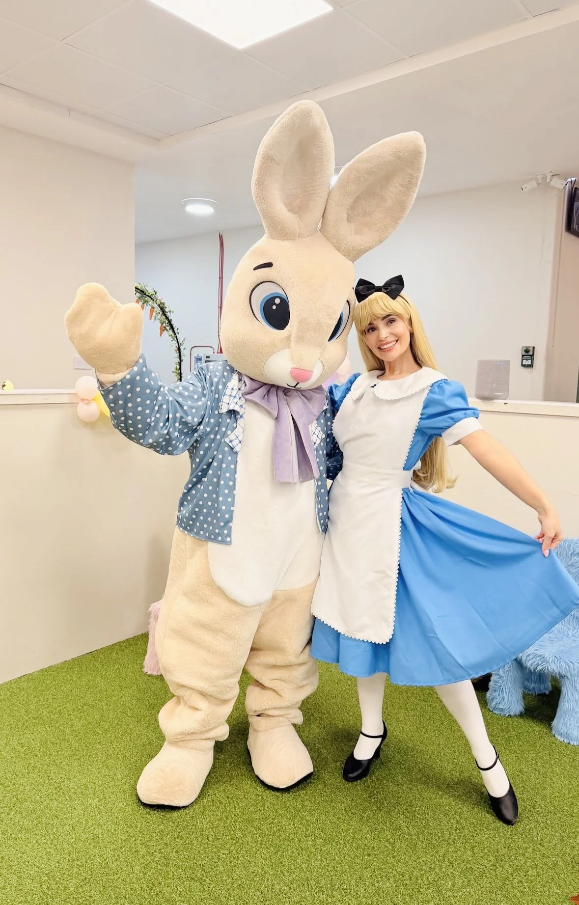A person dressed in a large bunny costume with big blue eyes, wearing a polka dot shirt and a purple bow, standing next to a woman in a blue and white dress with a black bow in her hair, both smiling and posing for the photo.