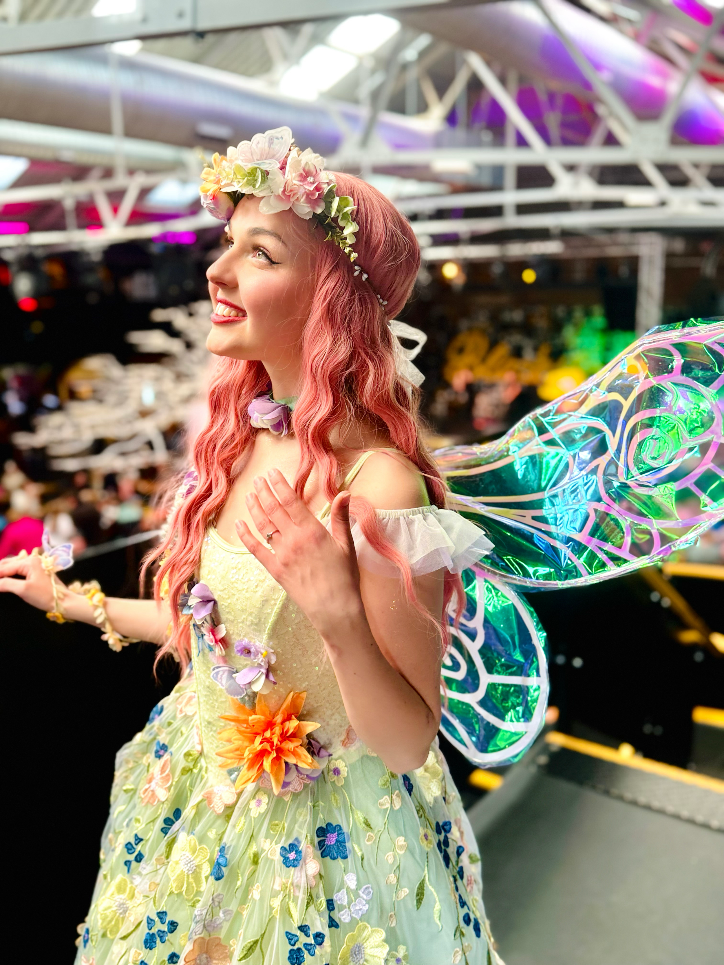 A woman dressed as a fairy or princess in a floral dress with butterfly wings and a floral headband, smiling at an indoor event.