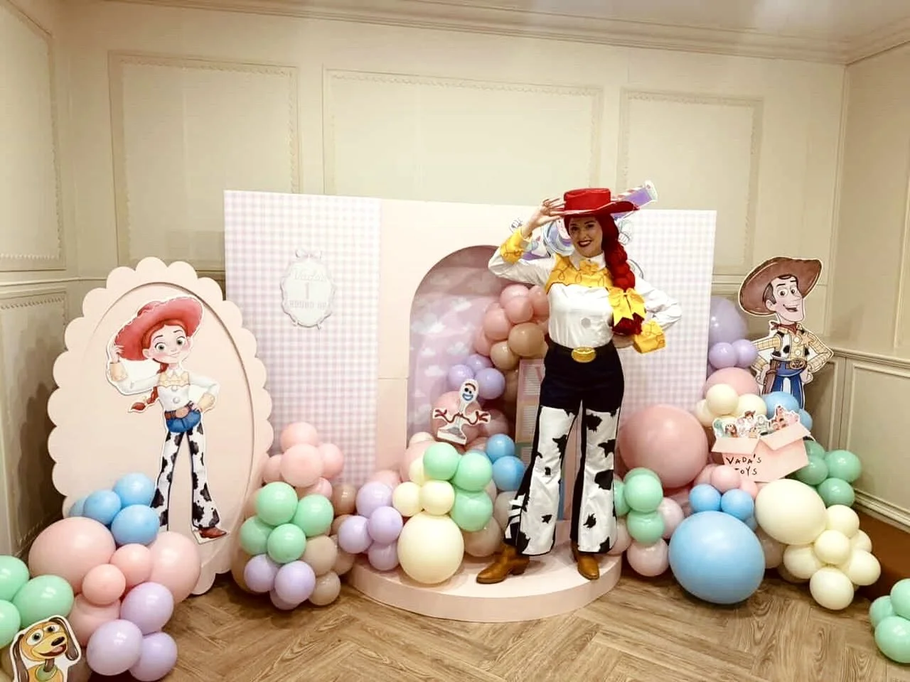 Decorated party scene with life-sized Buzz Lightyear character from Toy Story, surrounded by pastel-colored balloons and cutouts of Jessie and Woody characters, in a room with light-colored walls.