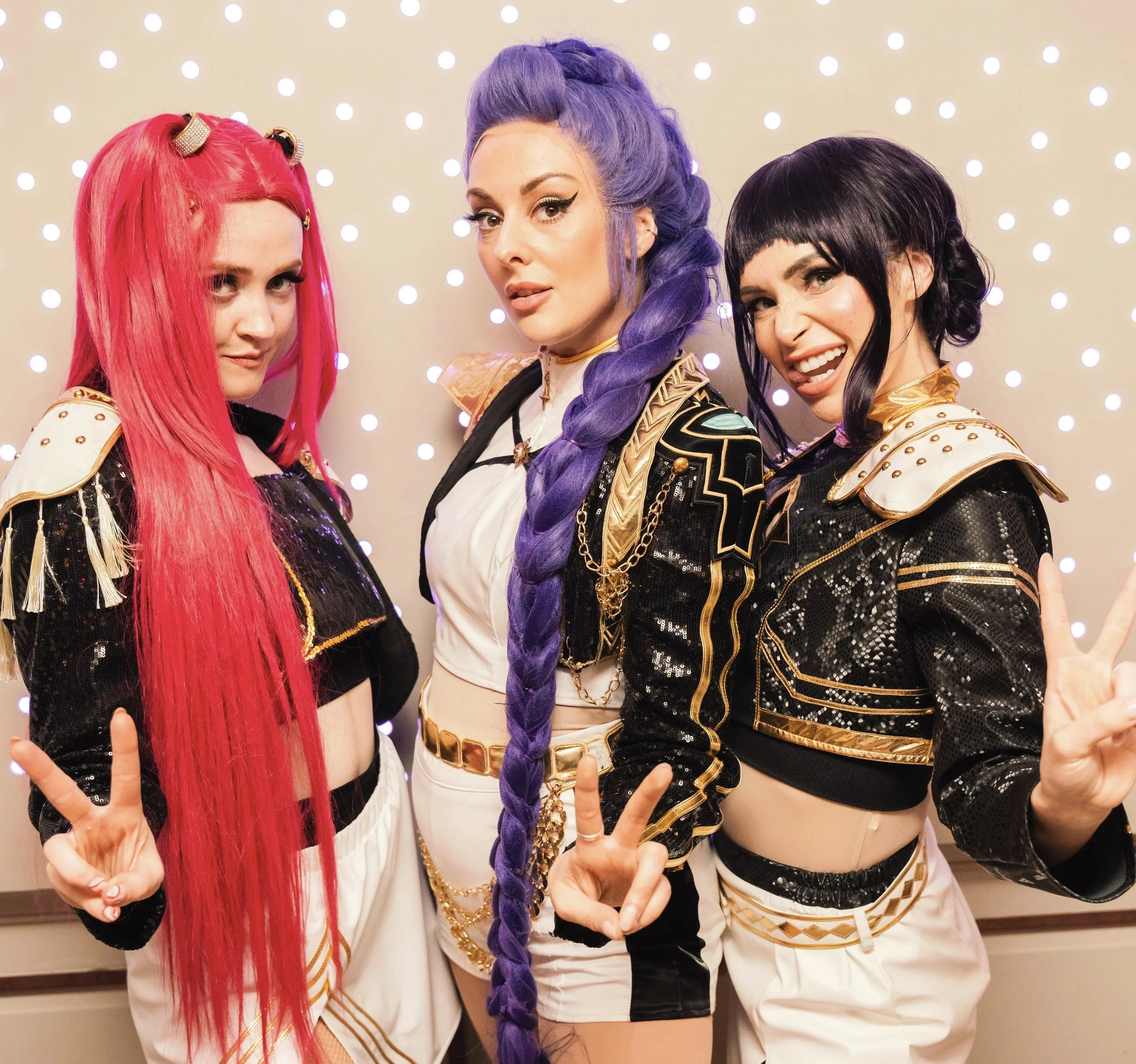 Three women dressed in futuristic, shiny black and gold costumes with colorful wigs posing against a background of bright lights.