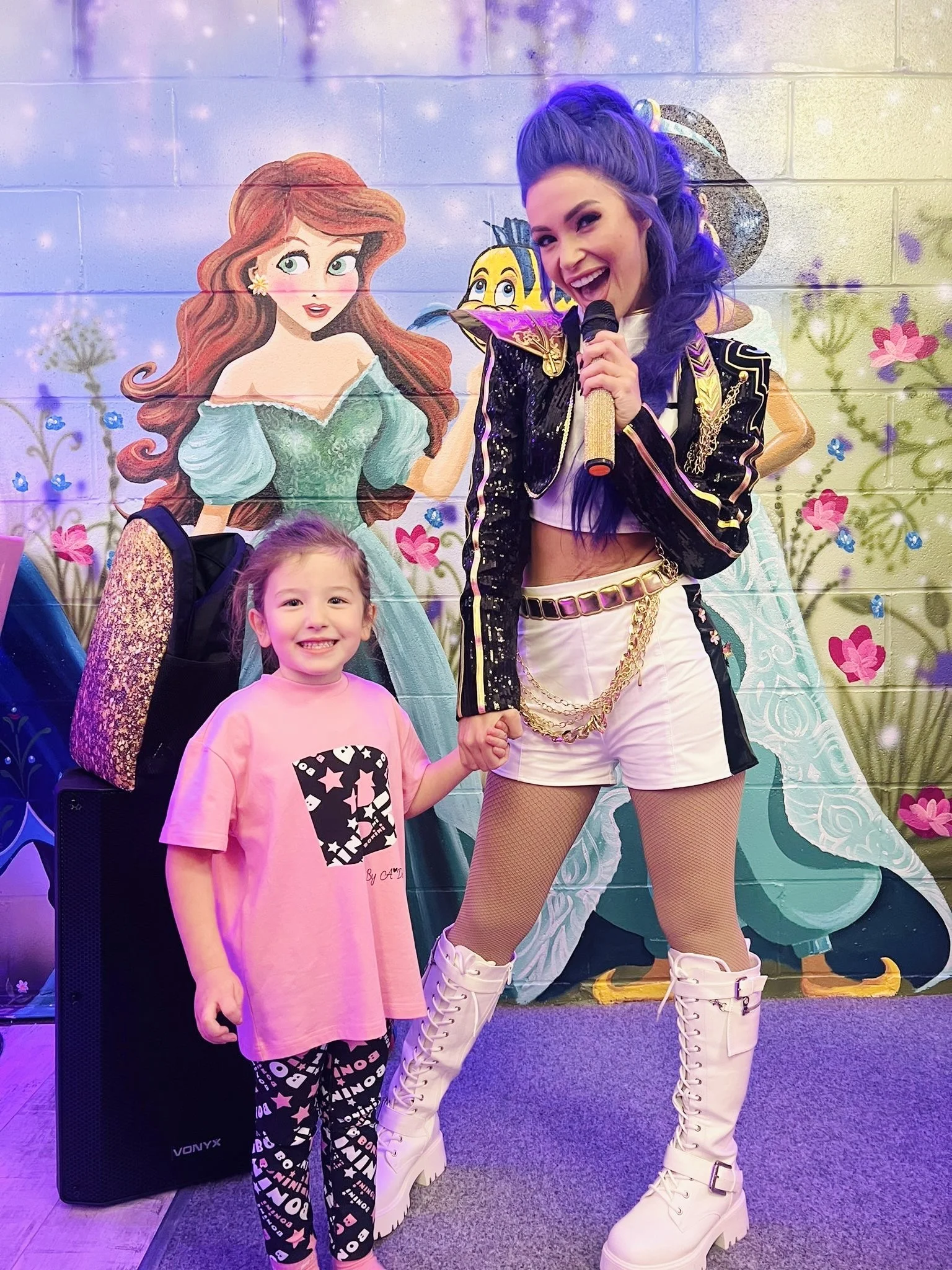 A woman with purple hair in a colorful outfit holding a microphone, standing next to a young girl in a pink dress with star and letter patterns holding hands, in front of a mural of Disney characters Ariel and Flounder.