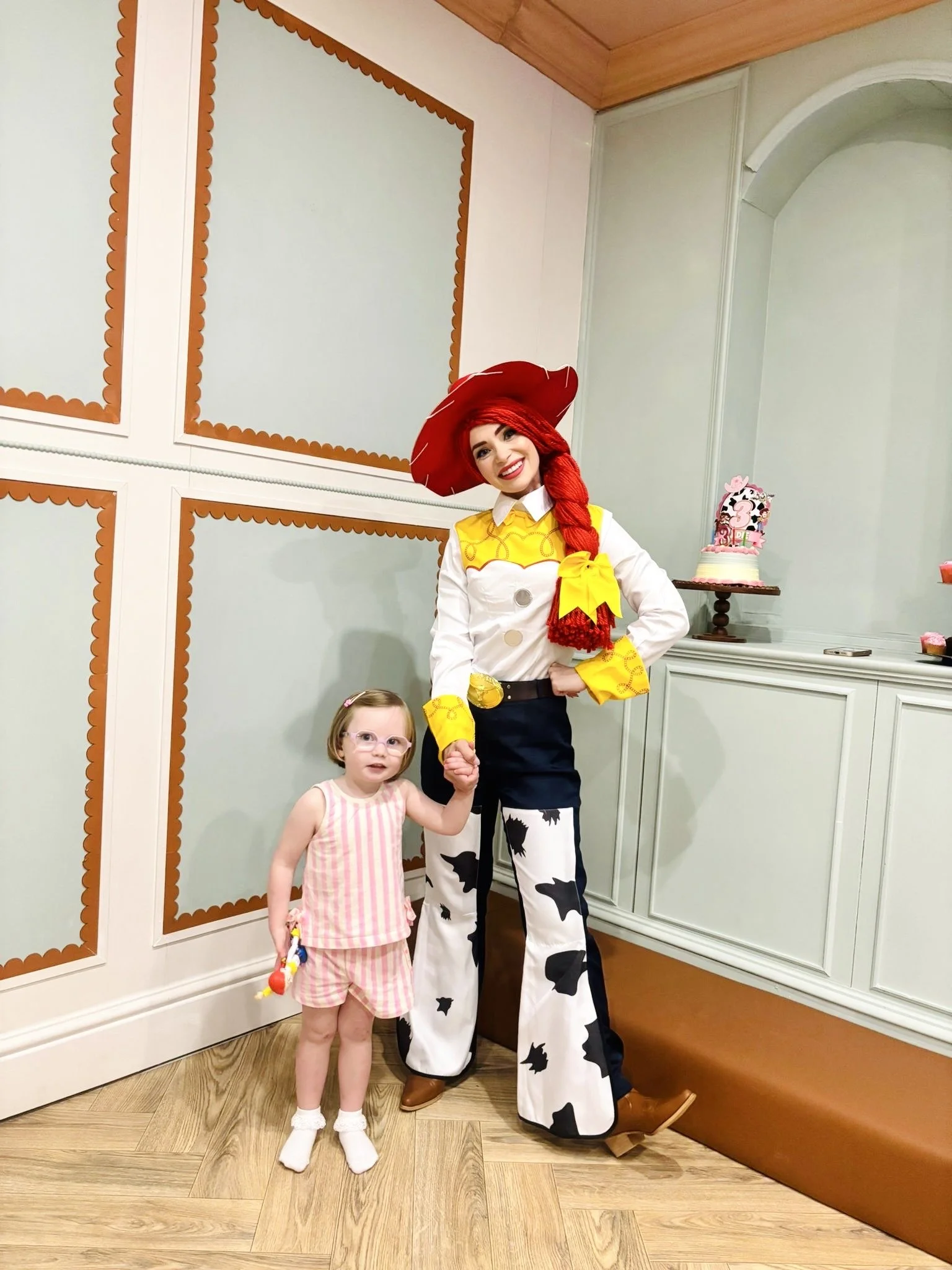A woman dressed as a cow mascot holding hands with a young girl at a birthday celebration. The woman is smiling and wearing a red hat and a costume with black and white cow spots. The young girl, wearing glasses and pink pajamas, holds a party favor.