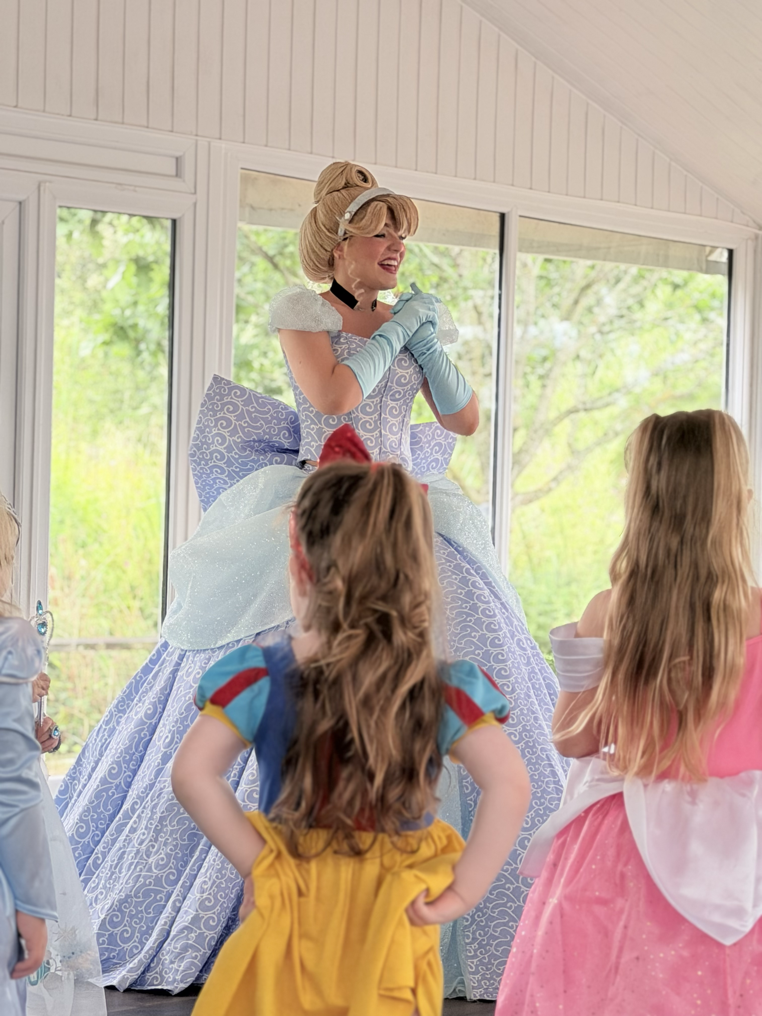 A woman dressed as a princess, wearing a blue gown with white lace and gloves, is smiling and holding her hands near her chest at a children's party. Children in costumes are gathered around, with colorful decorations and large windows showing trees 
