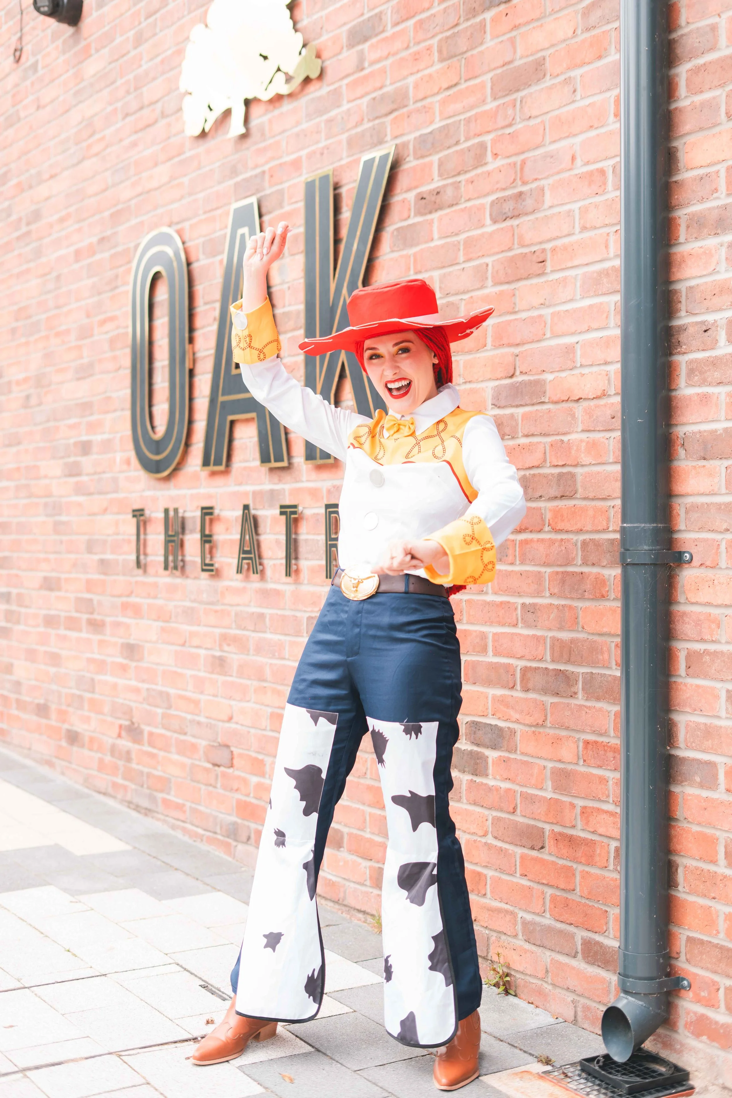 Woman in cow print pants, cowboy boots, yellow and white shirt, and red hat posing in front of a brick wall with large black letters that spell out 'OKAY THEATER' behind her. She is smiling and making a gesture with her right hand.