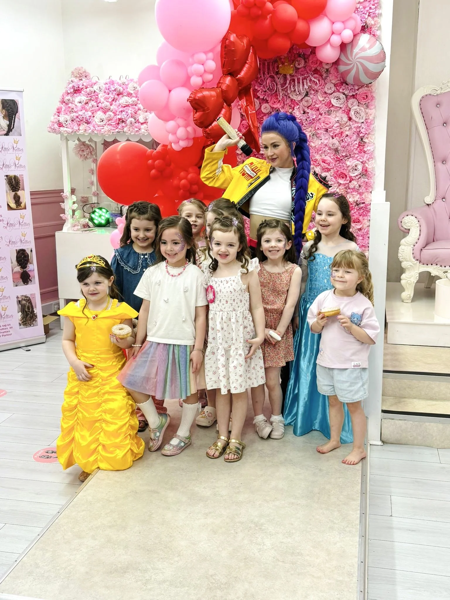 A group of young girls and a woman with bright blue braided hair posing together indoors for a photo at a celebration with pink and red balloons and a pink floral backdrop.