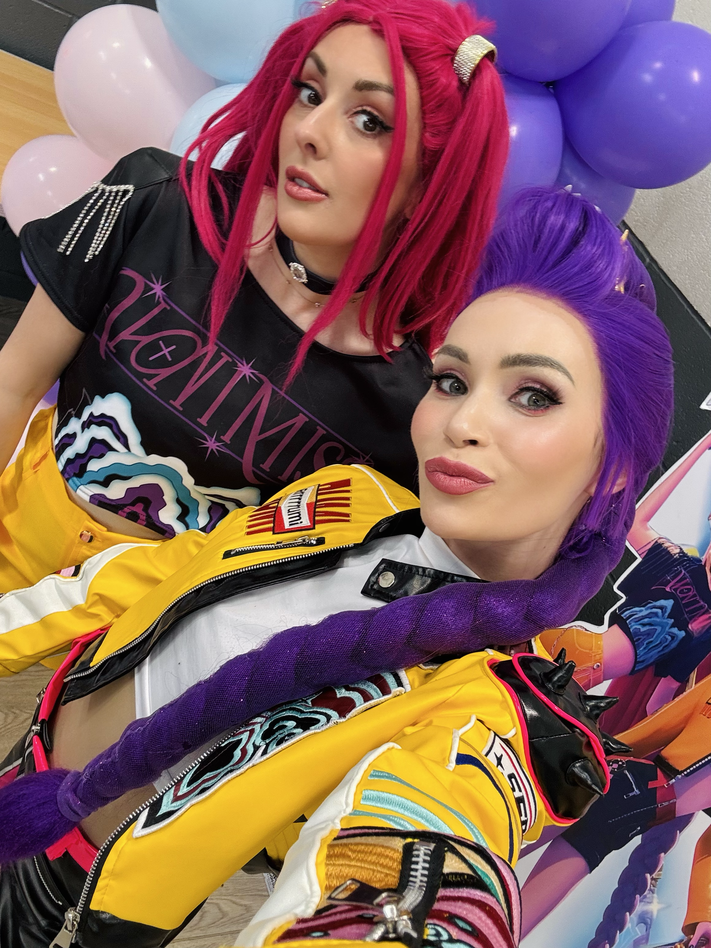 Two women with brightly colored pink and purple hair taking a close-up selfie. They are both wearing sporty outfits and standing against a background of purple and pink balloons.