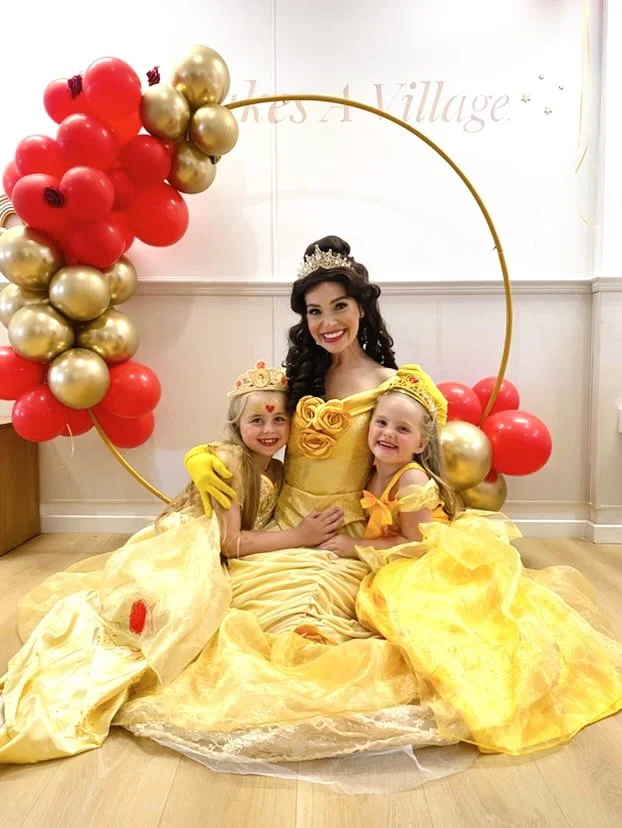 Three females dressed as princesses, two young girls and one adult woman, pose together smiling with a balloon backdrop featuring red and gold balloons forming a semi-circle.