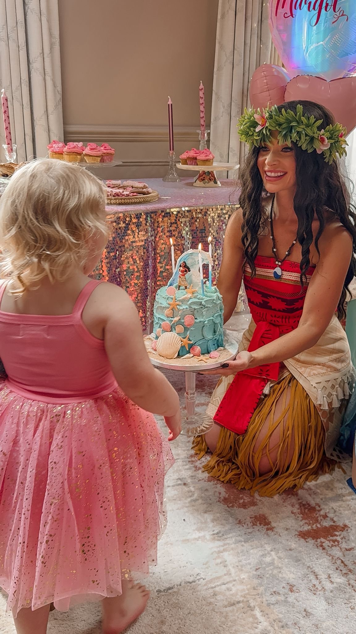 A woman wearing a floral crown and colorful dress holding a cake, with a young girl in a pink dress in front of her. In the background, a table with pink cupcakes, candles, and balloons, including a large helium balloon with the word 'HAPPY' on it.