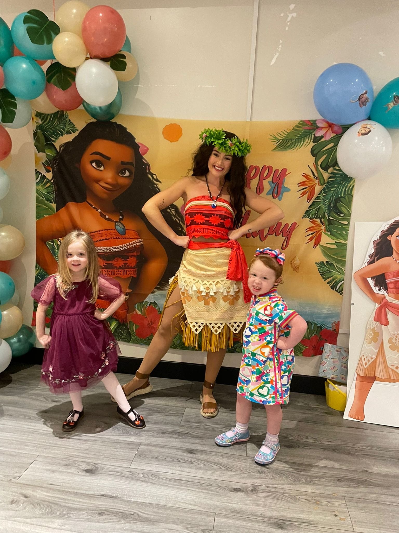 A woman dressed in a tropical outfit, wearing a leafy crown, posing with two young girls in colorful party clothes at a celebration with a Hawaii theme, surrounded by balloons and a tropical backdrop.