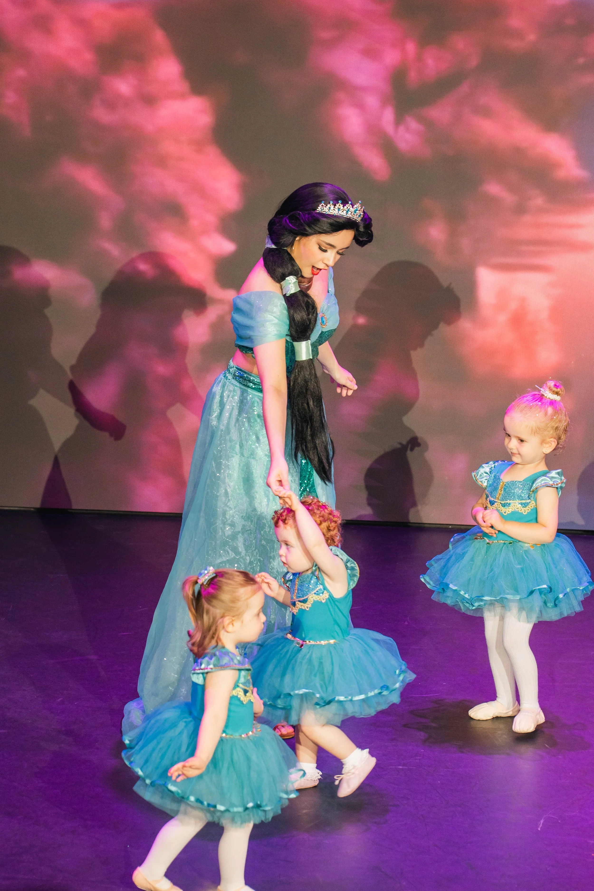 A woman dressed as a princess, wearing a tiara and a flowing blue gown, is holding a child's hand during a ballet recital. Several young girls in blue tutus and ballet slippers are dancing on stage. The background features pink and purple lighting wi