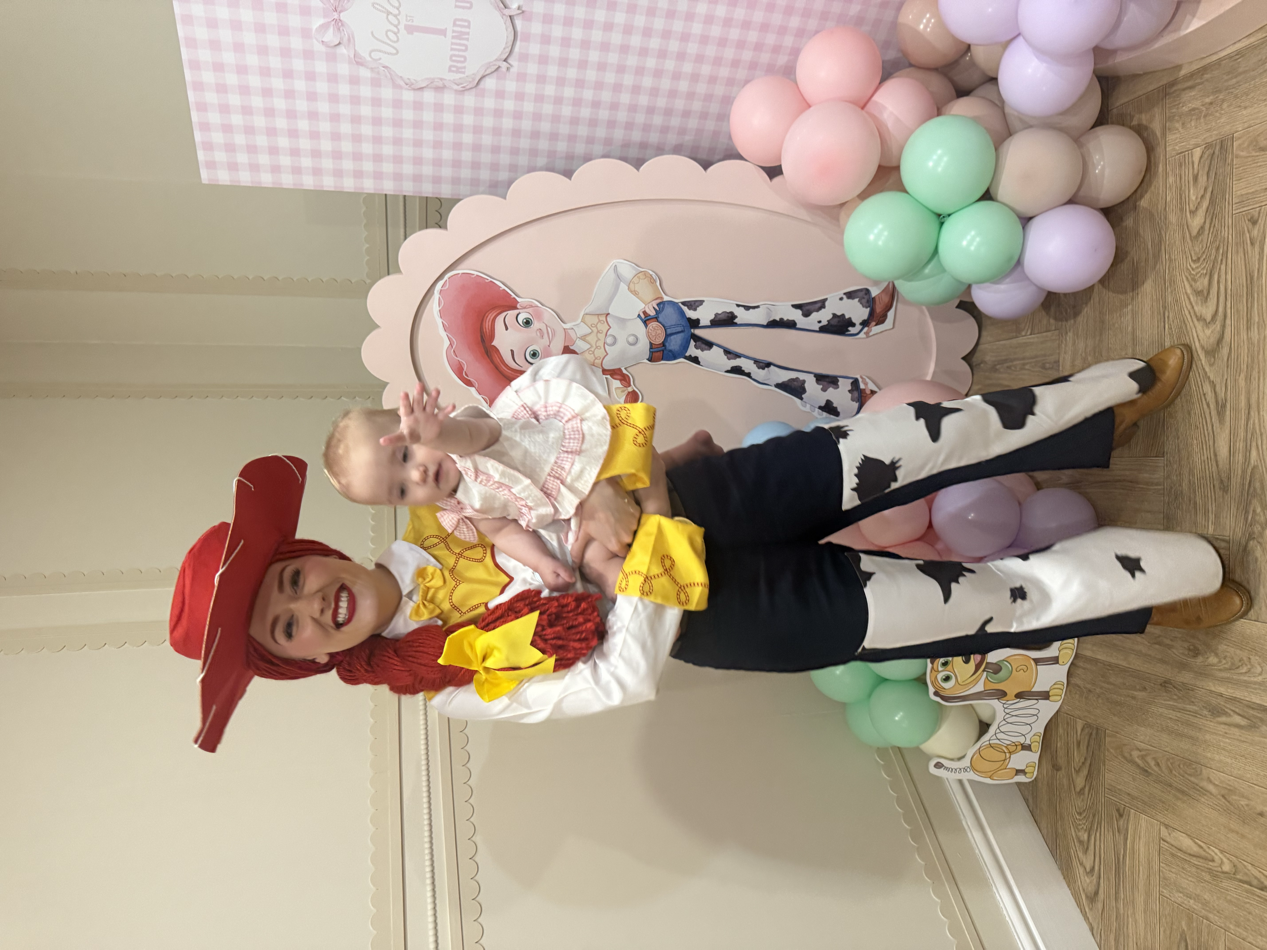 A young girl dressed as Jessie from Toy Story and a woman dressed as Jessie from Toy Story at a birthday party. They are standing next to a decorative backdrop with pastel-colored balloons, featuring a cartoon image of Jessie. The woman is wearing a 
