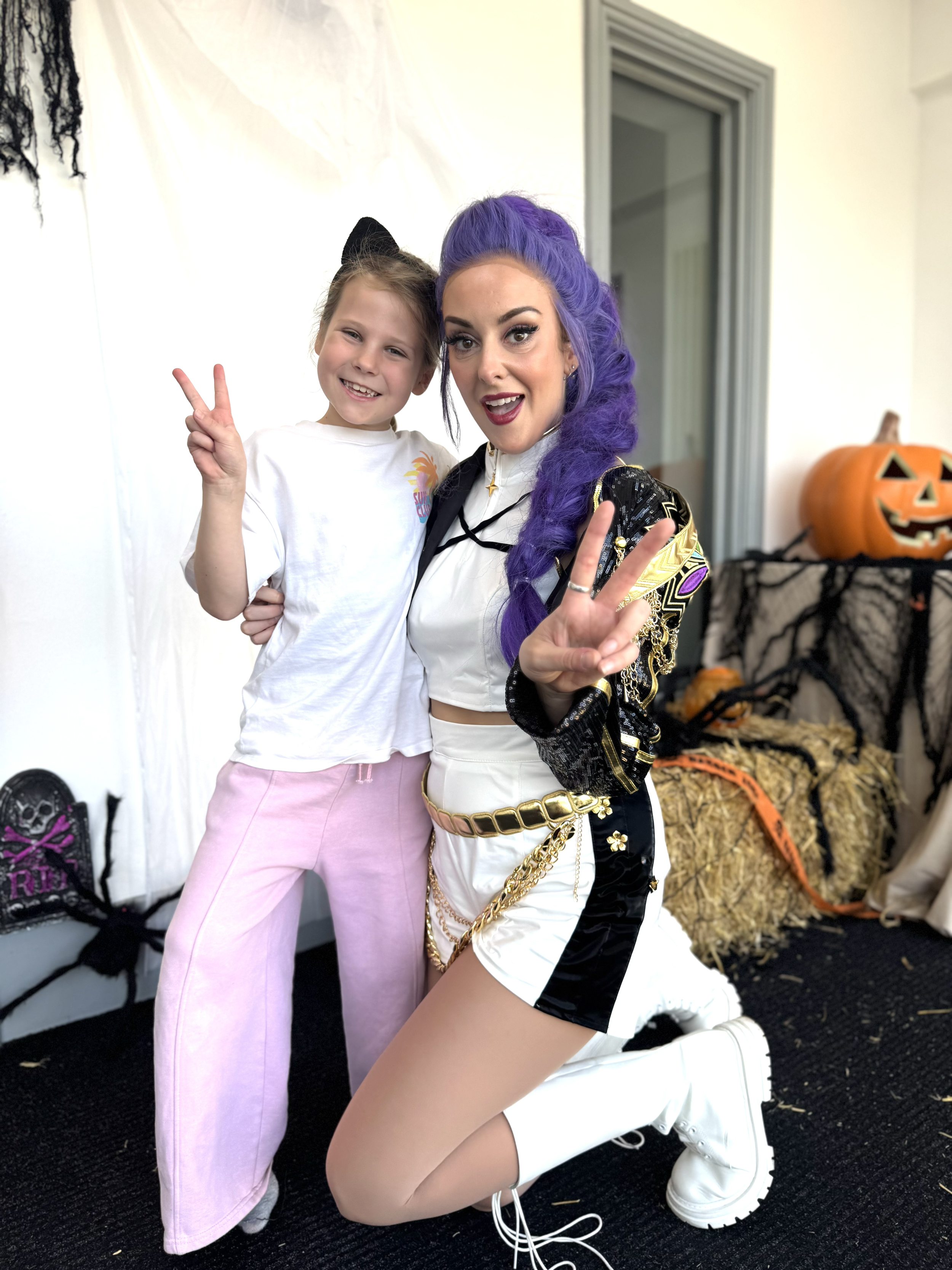 A young girl wearing a white t-shirt with a colorful sun graphic and pink pants, smiling and making a peace sign with her right hand, standing next to a woman with purple hair styled in a large wave, wearing a shiny black and gold jacket, white short