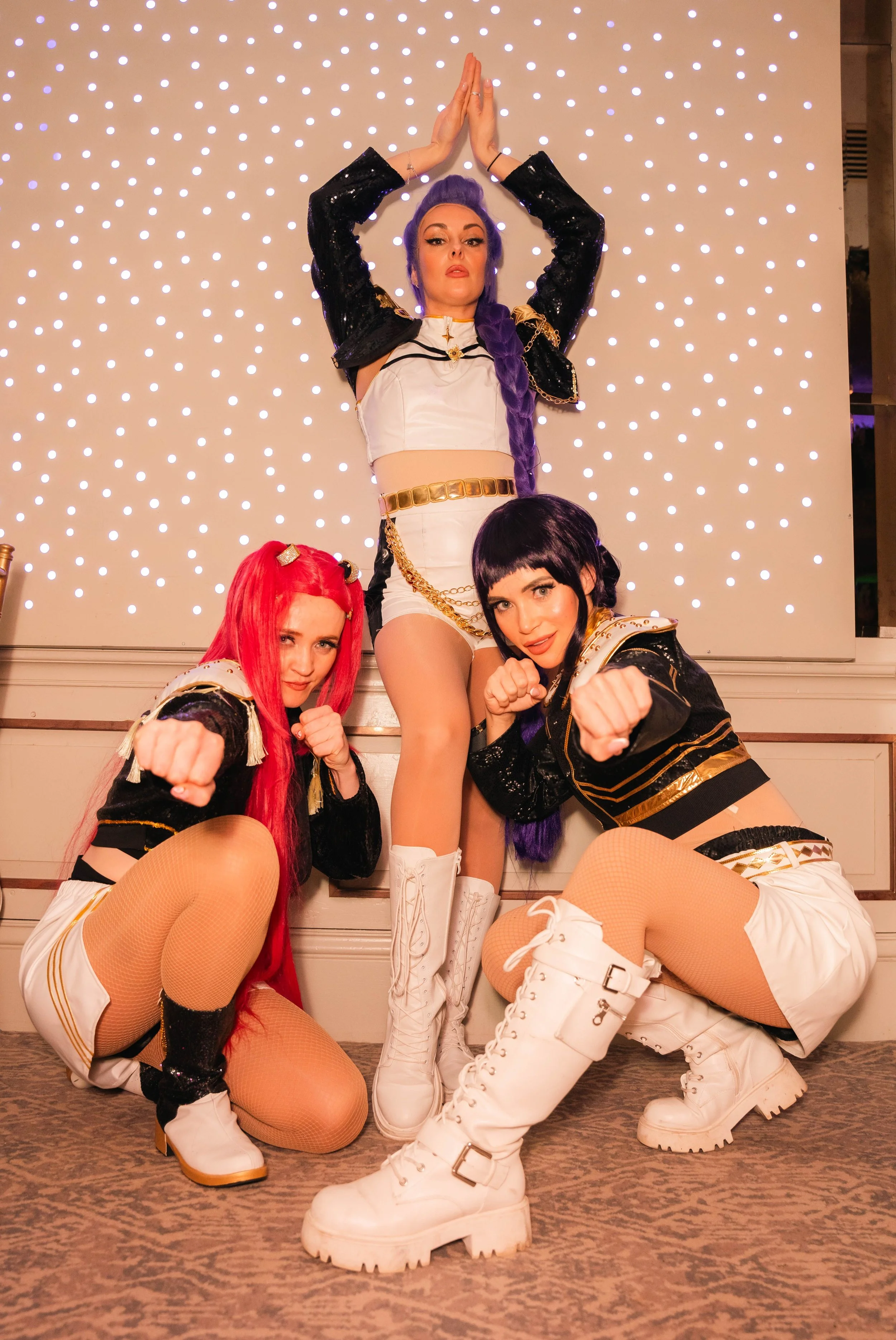Four women in cosplay costumes posing in front of a lit-up background. The woman in the center stands with arms raised, wearing a white and black outfit with gold accents and white boots. The other three women squat in front, dressed in similar outfi