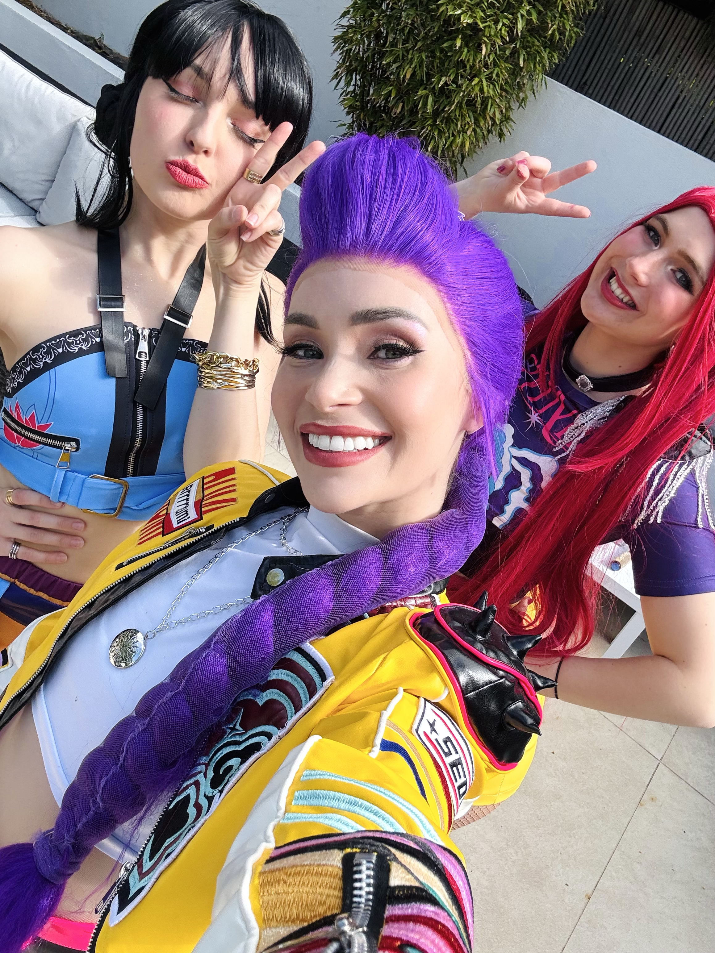 Three women taking a selfie together; one woman with black hair and red lipstick, one with purple hair, and one with red hair, all smiling and making peace signs.