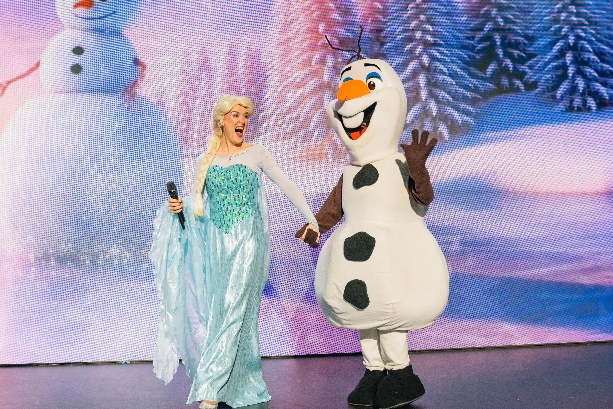 Elsa from Frozen and a person dressed as Olaf from Frozen on stage, smiling and waving.